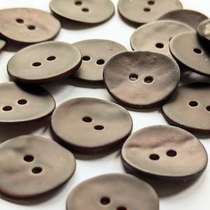 Puede incluir: Una colección de botones marrones de dos agujeros. Los botones están hechos de un material natural y tienen una superficie ligeramente irregular.