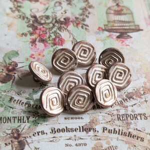 Puede incluir: Una colección de diez botones de metal de color dorado con un diseño en espiral. Los botones están dispuestos en semicírculo sobre una superficie estampada.