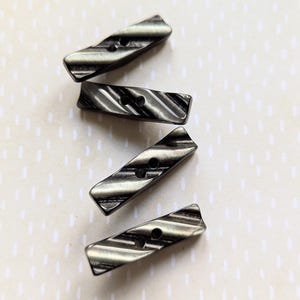 Puede incluir: Cuatro botones rectangulares de color gris oscuro con un patrón diagonal acanalado. Cada botón tiene dos agujeros para coser. Los botones tienen una forma ligeramente curvada y están dispuestos diagonalmente sobre un fondo claro.