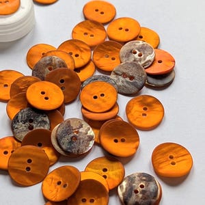 Può includere: Una collezione di bottoni arancioni e marroni, ciascuno con due fori. I bottoni variano per colore e consistenza, alcuni con un aspetto marmorizzato. Sono sparsi su una superficie bianca, suggerendo un progetto di artigianato o cucito.