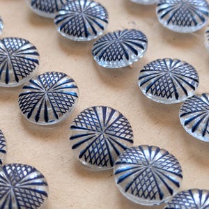 May include: A collection of clear, round glass beads with a detailed blue and silver geometric design. The beads have a domed shape and are scattered across a neutral surface, showcasing their intricate patterns.