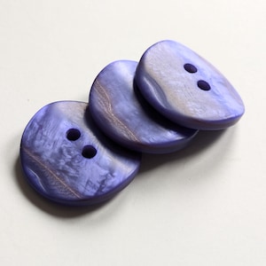 Puede incluir: Tres botones iridiscentes de color morado con dos agujeros cada uno. Los botones están apilados uno encima del otro.