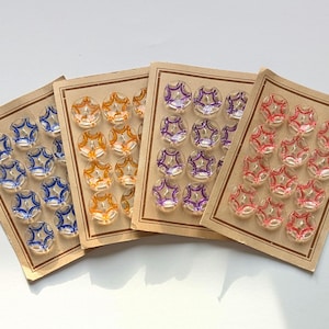 Könnte beinhalten: Vier Sets Knöpfe im Vintage-Stil auf Karton. Jedes Set hat eine andere Farbe: Blau, Orange, Lila und Rot. Die Knöpfe haben ein klares, sternförmiges Design mit farbigen Akzenten, ideal zum Nähen oder für Bastelprojekte.