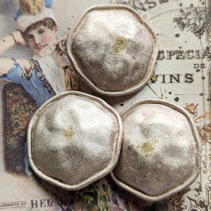 Puede incluir: Tres botones octogonales de color plateado con una superficie ligeramente elevada y texturizada. Los botones tienen un aspecto desgastado y envejecido con un toque de oro.