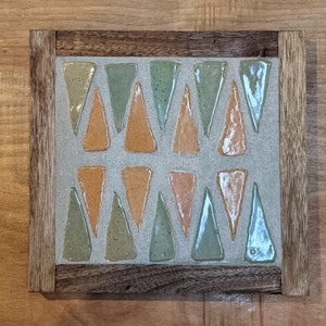 Può includere: Una cornice in legno con uno sfondo grigio e un motivo geometrico di piastrelle triangolari in tonalità di verde, arancione e marrone.