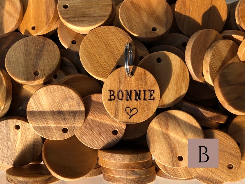 May include: A close-up of a pile of round wooden tags with a single tag engraved with the name "BONNIE" and a heart. The tags have a hole for a key ring.