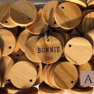 May include: A close-up of a pile of unfinished wooden dog tags. One tag is engraved with the name "BONNIE" and a bone symbol. The letter "A" is visible in the lower right corner of the image.