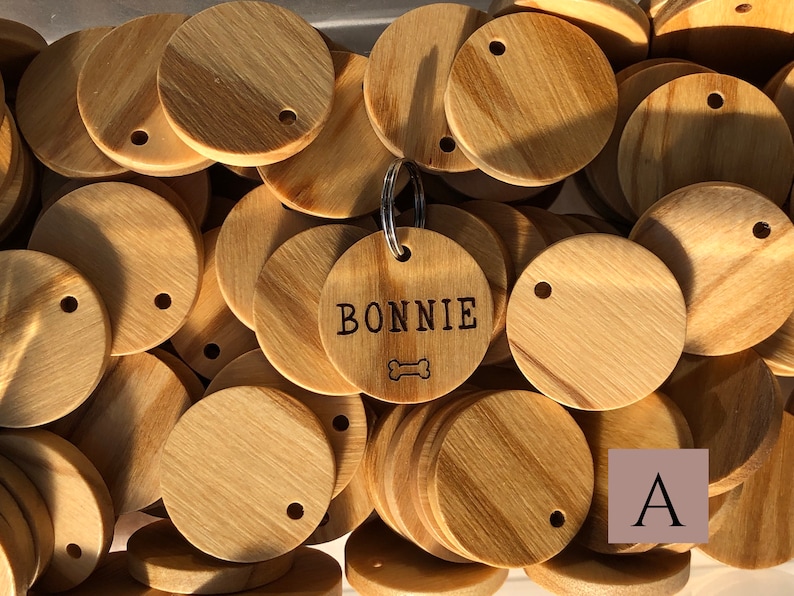 May include: A close-up of a pile of unfinished wooden dog tags. One tag is engraved with the name "BONNIE" and a bone symbol. The letter "A" is visible in the lower right corner of the image.