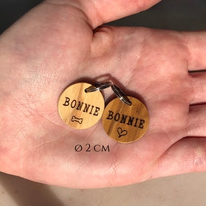 May include: Two wooden dog tags with the name "BONNIE" engraved on them. One tag has a bone symbol and the other has a heart symbol. The tags are 2 cm in diameter.