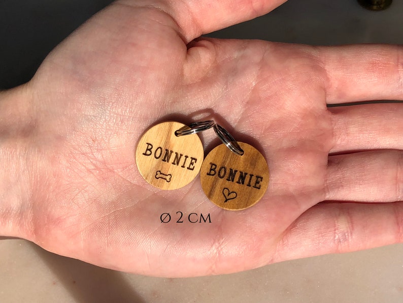 May include: Two wooden dog tags with the name "BONNIE" engraved on them. One tag has a bone symbol and the other has a heart symbol. The tags are 2 cm in diameter.