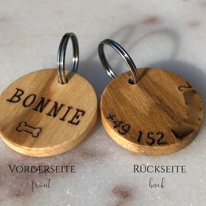 May include: A wooden dog tag with a metal ring. The front side is engraved with the name "BONNIE" and a bone symbol. The back side is engraved with the phone number "+49 152".