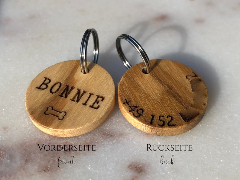 May include: A wooden dog tag with a metal ring. The front side is engraved with the name "BONNIE" and a bone symbol. The back side is engraved with the phone number "+49 152".