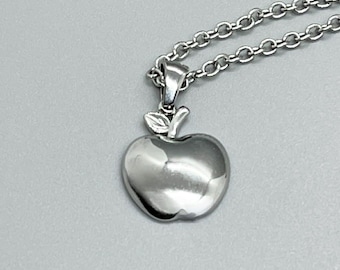 May include: A silver-colored apple pendant necklace. The pendant is a detailed apple shape with a small leaf. The chain is a delicate link style. The necklace is displayed against a gray background.