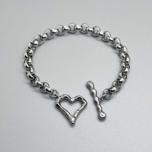 May include: A silver chain bracelet with a heart-shaped charm. The heart charm is open and has a rough, textured surface.