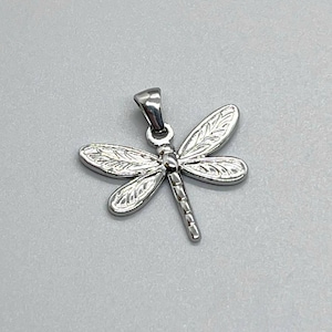May include: A silver dragonfly pendant with detailed wing patterns. The pendant has a loop at the top for a chain. The dragonfly's body is segmented, and the wings are textured with vein-like designs. The pendant is set against a light gray background.