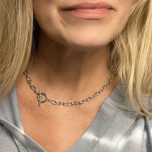 May include: A silver chain necklace with a toggle clasp. The necklace is worn by a person with blonde hair.