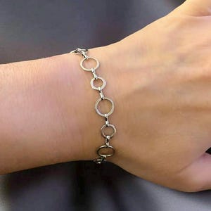 May include: A silver bracelet featuring a series of interconnected circles of varying sizes. The bracelet is displayed on a wrist, showcasing its design and how it lays. The jewelry is simple and elegant, suitable for everyday wear.