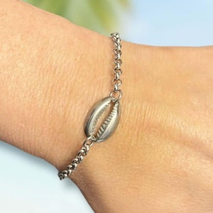May include: A silver chain bracelet with a seashell charm. The bracelet is on a person's wrist.