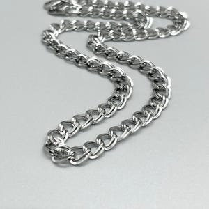 May include: A silver-toned double curb chain necklace featuring interlocking oval links with a polished, reflective metallic finish. The chain is arranged in a curved, flowing pattern, showcasing the uniform structure and smooth texture of the metal links.