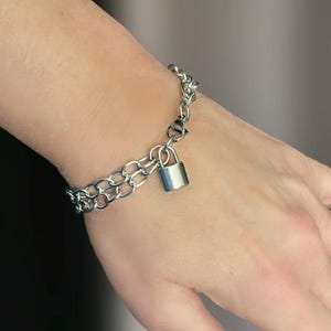 May include: A silver chain bracelet with a padlock charm. The bracelet is made of two chains, one thicker than the other, and the padlock is attached to the thinner chain.