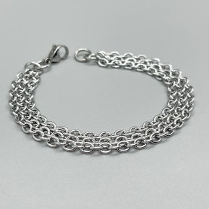 May include: A silver-colored bracelet with three parallel chains of small, round links. The bracelet has a clasp closure and is displayed on a light gray background. The jewelry is made of a shiny, metallic material.