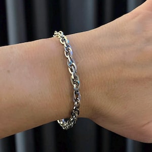 May include: A silver-colored chain bracelet with an oval link design. The bracelet is worn on a wrist, showcasing the polished metal and the way it catches the light. The jewelry is a simple yet elegant accessory.