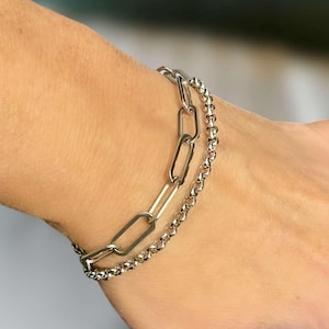 May include: Two silver chain bracelets stacked on a person's wrist. The top bracelet has larger, oval-shaped links. The bottom bracelet has smaller, round links.