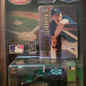 May include: A green toy truck with a Milwaukee Brewers logo and the name "Jeromy Burnitz" on the side. The truck is on a white background with a baseball field and a baseball card in the background.