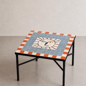 May include: A square coffee table with a black metal frame and a tiled tabletop. The surface features a central sun-like face motif in white, surrounded by a blue grid border and an outer edge of alternating orange and white rectangular tiles.
