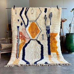 Peut inclure: Un grand tapis crème avec un motif géométrique. Le tapis présente des formes orange, bleu marine et roses. Le tapis est suspendu à une barre métallique, mettant en valeur sa texture et son design. Le tapis a une bordure à franges.
