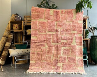 Puede incluir: Una alfombra de color rosa y crema con un diseño geométrico. La alfombra tiene un borde de flecos y está hecha de un material suave y esponjoso.