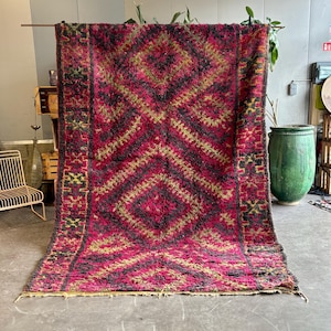 May include: A large, rectangular rug with a geometric diamond pattern in shades of red, black, and gold. The rug is displayed vertically, showcasing its intricate design and textured surface. The rug's dimensions are not visible.