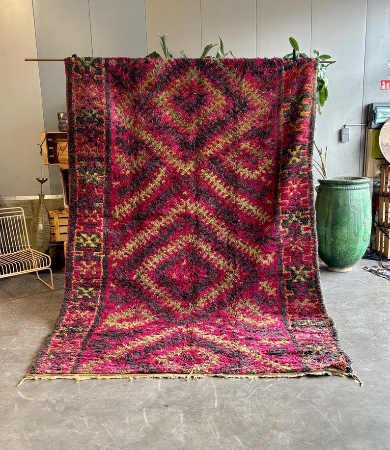 May include: A large, rectangular rug with a geometric diamond pattern in shades of red, black, and gold. The rug is displayed vertically, showcasing its intricate design and textured surface. The rug's dimensions are not visible.