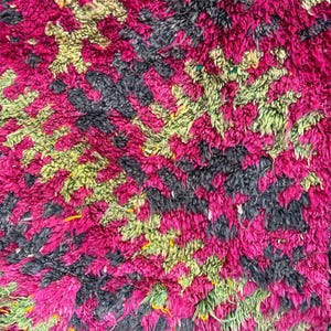 May include: Close-up of a plush rug with a vibrant, textured design. The rug features a pattern of magenta, black, and light green shapes. The thick pile creates a soft, inviting appearance, suitable for home decor.