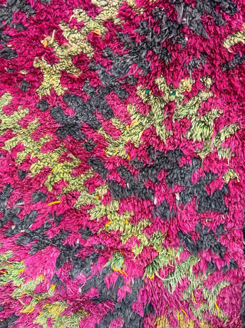 May include: Close-up of a plush rug with a vibrant, textured design. The rug features a pattern of magenta, black, and light green shapes. The thick pile creates a soft, inviting appearance, suitable for home decor.