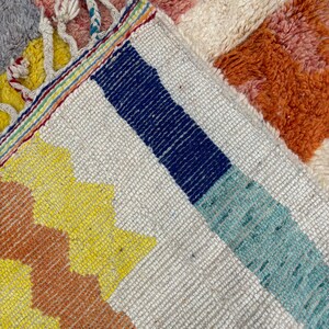 May include: Close-up of a colorful woven rug with a geometric pattern. The rug features stripes of blue, teal, yellow, and orange against a white background. The rug has a fringe border.