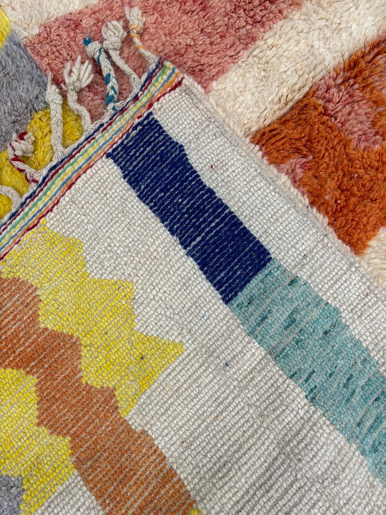May include: Close-up of a colorful woven rug with a geometric pattern. The rug features stripes of blue, teal, yellow, and orange against a white background. The rug has a fringe border.