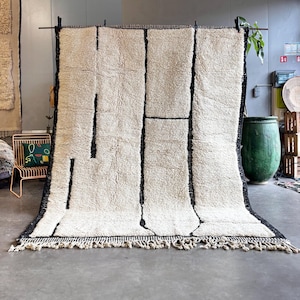 May include: A large, rectangular rug with a cream-colored background and bold black geometric lines. The rug has a thick, textured pile and a fringe along the bottom edge. The rug is displayed indoors, likely for home decor.