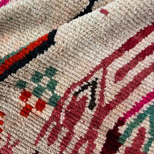 May include: A close-up of a colourful Moroccan rug with a cream background and intricate patterns in shades of red, pink, green, and black.