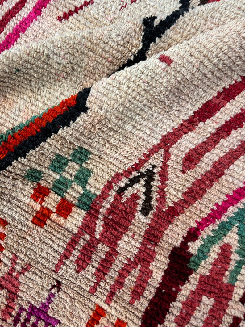 May include: A close-up of a colourful Moroccan rug with a cream background and intricate patterns in shades of red, pink, green, and black.