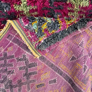 May include: A close-up of a handmade rug with a vibrant color palette. The rug features a mix of textures and patterns, including a plush, shaggy top layer in shades of fuchsia, green, and charcoal. The lower layer displays a geometric design in purple and yellow.