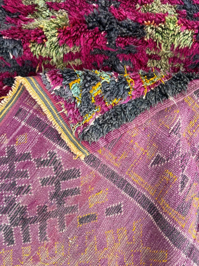 May include: A close-up of a handmade rug with a vibrant color palette. The rug features a mix of textures and patterns, including a plush, shaggy top layer in shades of fuchsia, green, and charcoal. The lower layer displays a geometric design in purple and yellow.