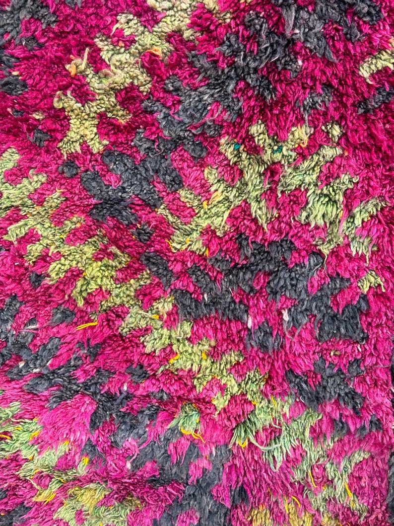 May include: Close-up of a plush rug with a vibrant, textured design. The rug features a pattern of magenta, black, and light green shapes. The thick pile creates a soft, inviting appearance, suitable for home decor.