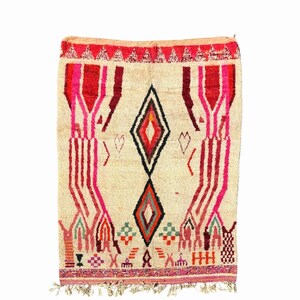 May include: A beige rug with a geometric pattern in shades of pink, red, black, and green. The rug has a diamond shape in the centre and fringe on the bottom.