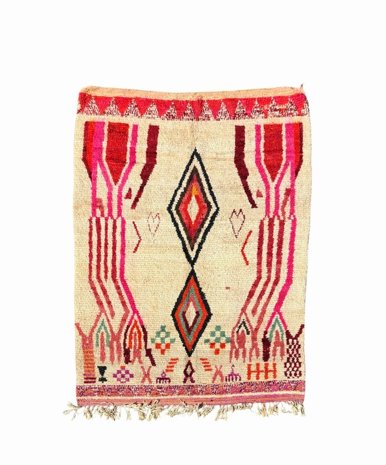 May include: A beige rug with a geometric pattern in shades of pink, red, black, and green. The rug has a diamond shape in the centre and fringe on the bottom.