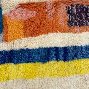 May include: A close-up of a colorful, striped rug with a fuzzy texture. The rug features shades of orange, yellow, blue, and white.