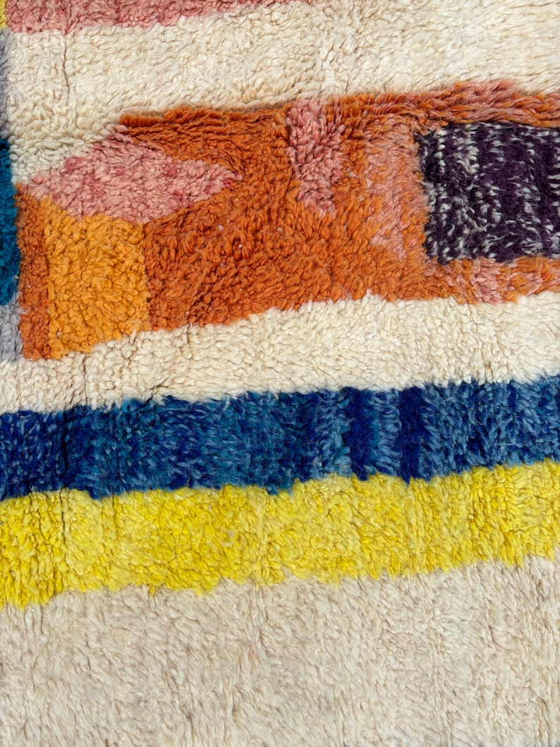 May include: A close-up of a colorful, striped rug with a fuzzy texture. The rug features shades of orange, yellow, blue, and white.