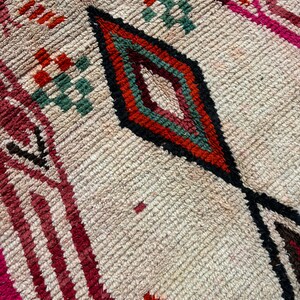 May include: Close-up of a beige rug with a diamond pattern in shades of red, orange, green, and black. The rug has a textured, woven surface.
