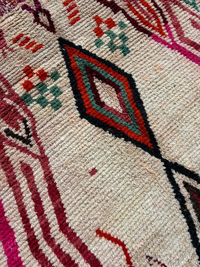 May include: Close-up of a beige rug with a diamond pattern in shades of red, orange, green, and black. The rug has a textured, woven surface.