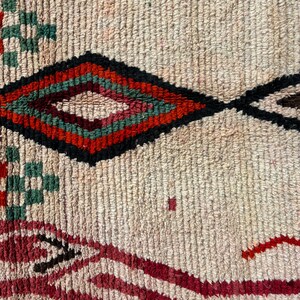 May include: Close-up of a woven rug with a diamond pattern in shades of red, green, and black on a cream background.